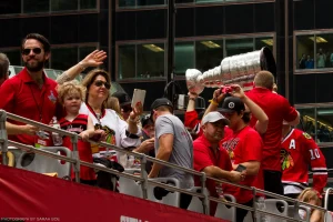 Brent Seabrook hoists the Stanley Cup on a bus with friends, family, and Patrick Sharp, while Chicagoans watch from adjacent buildings.